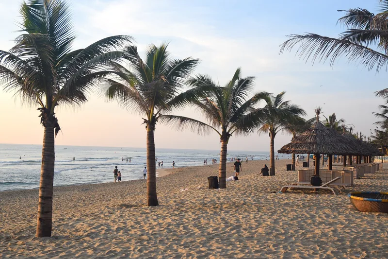 Palm trees line a sandy Da Nang beach with thatched umbrellas and people strolling at sunset.