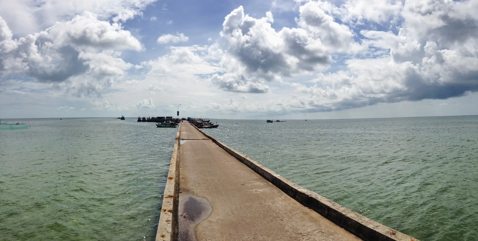 A long concrete pier stretches into the sea under a wide sky, with fishing boats near Ham Ninh, Phu Quoc.