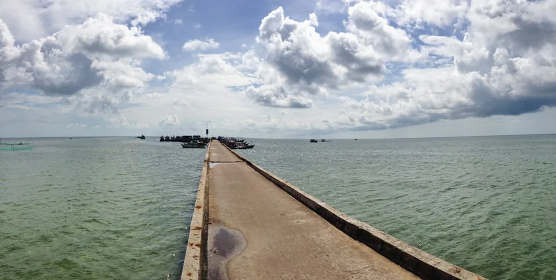 A long concrete pier stretches into the sea under a wide sky, with fishing boats near Ham Ninh, Phu Quoc.