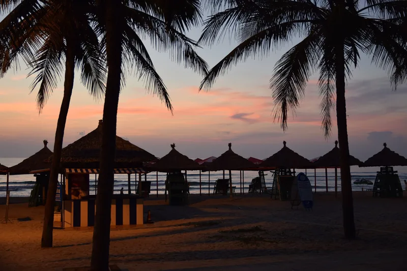 Palm trees and thatched beach umbrellas silhouetted against a colorful sunset sky on Da Nang beach.