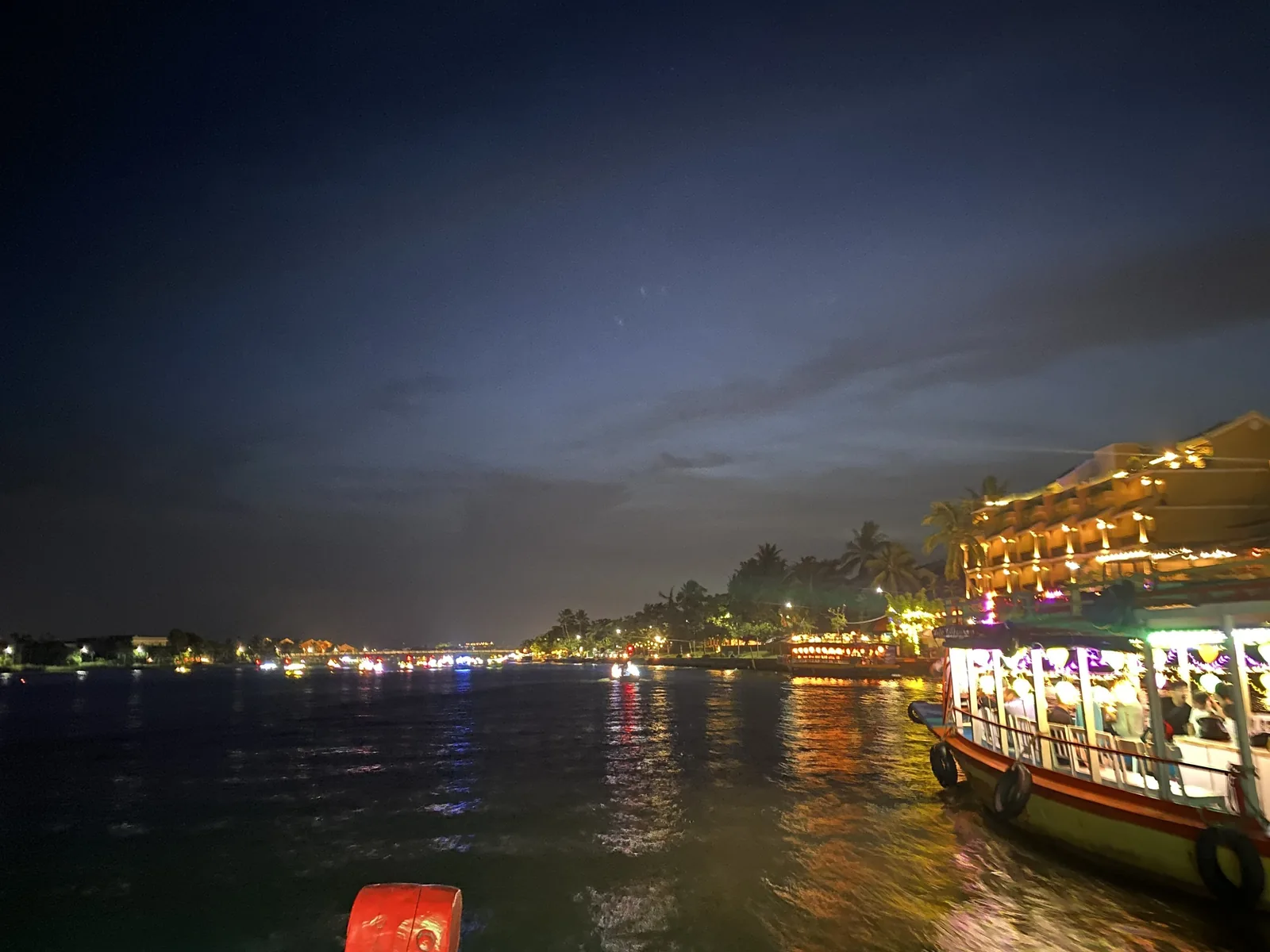 A lantern-lit tour boat glows on the Thu Bon River at night with Hoi An's riverside buildings reflecting on the water.