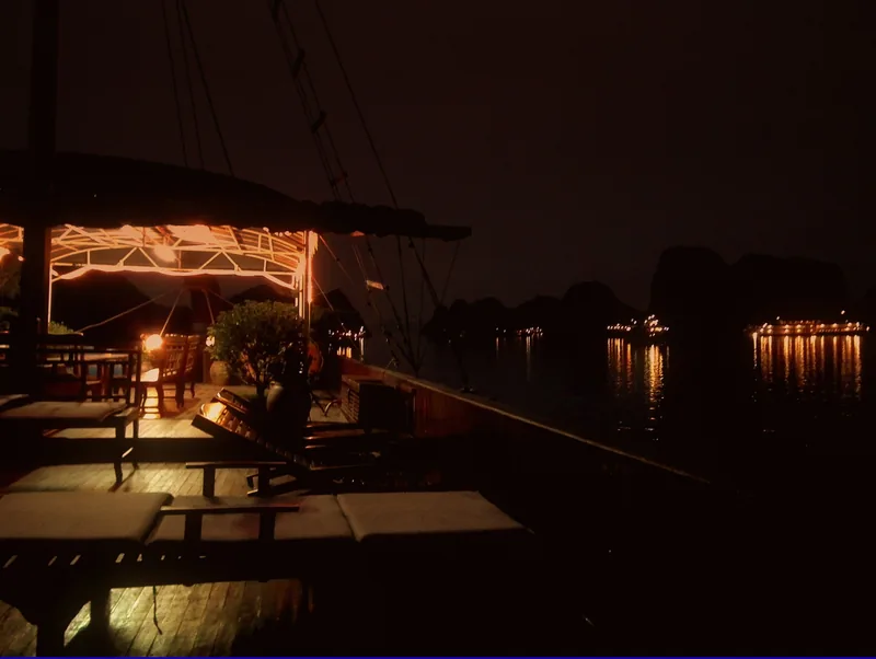 A cruise boat's lit deck with lounge chairs overlooks the dark waters of Ha Long Bay at night.