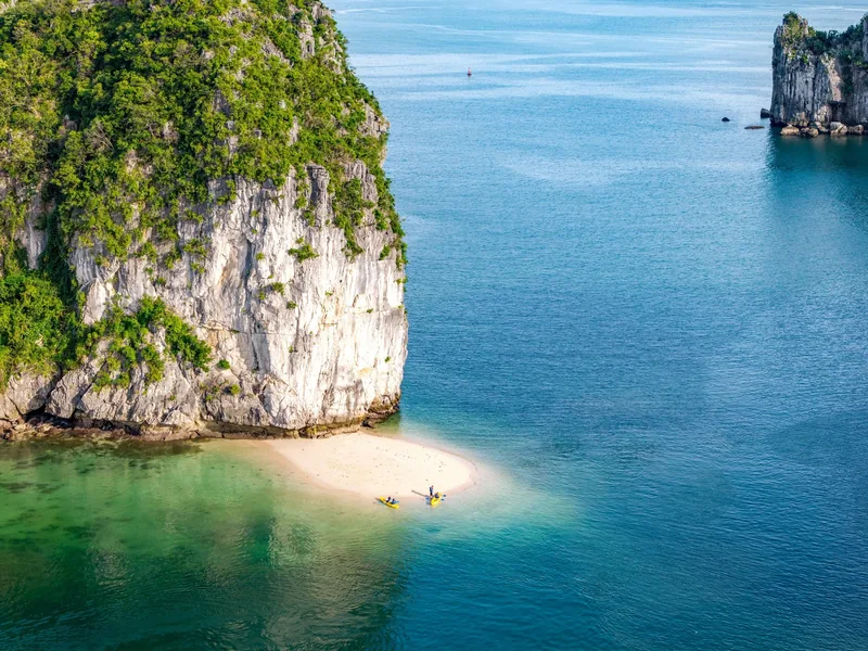 A yellow kayak rests on a small sandy beach beneath towering limestone cliffs in Lan Ha Bay.
