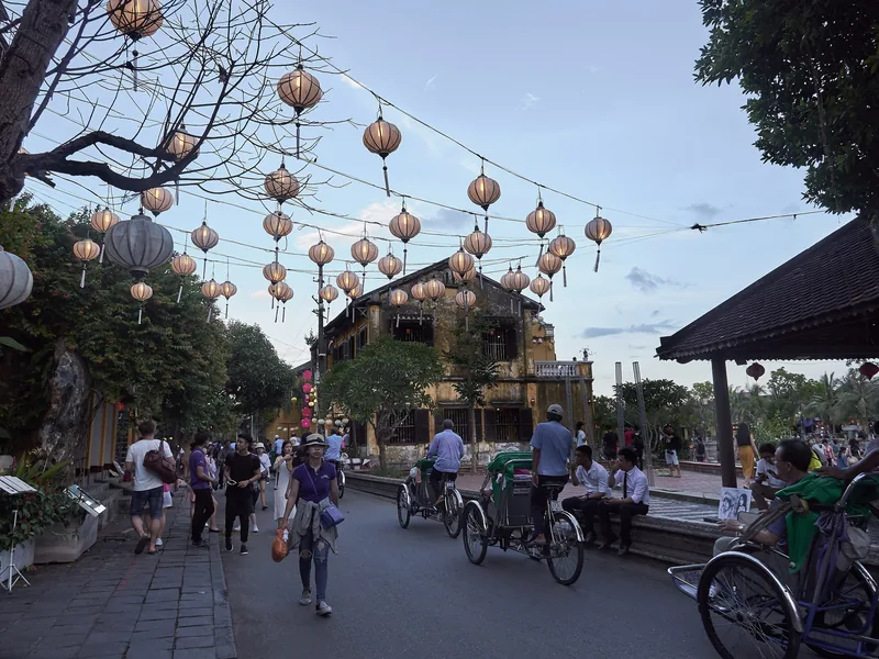 Silk lanterns hang over a lively street lined with yellow buildings as pedicabs and pedestrians pass at dusk in Hoi An.