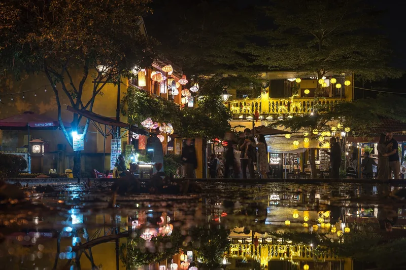 Glowing lanterns line riverside buildings in Hoi An's old town at night, reflected in the water below.