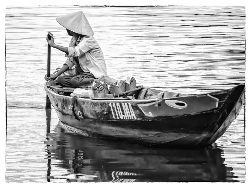 A person in a conical hat rows a small wooden boat on the calm Thu Bon River in Hoi An.