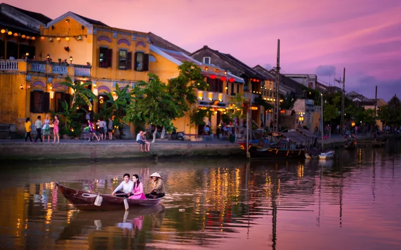 A boat glides along Hoi An's riverside at dusk, lined with yellow colonial buildings under a pink sky.