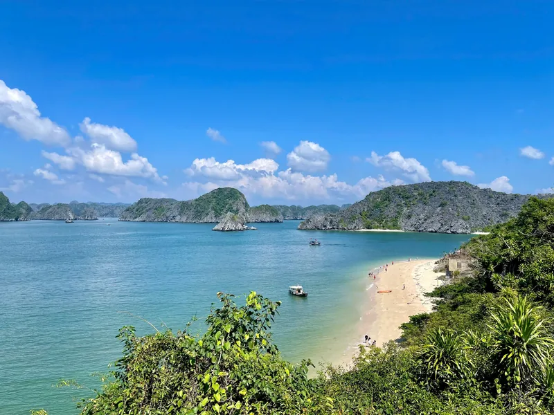 A crescent sandy beach curves along turquoise water surrounded by limestone karsts in Lan Ha Bay.