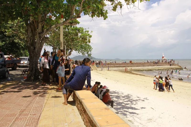 Visitors relax on a shaded seawall beside a sandy beach in Kep, Cambodia.