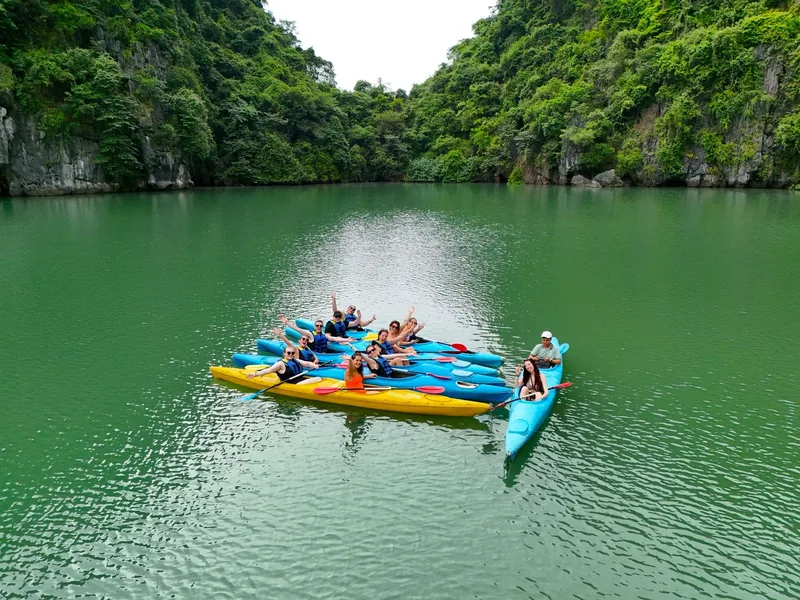 Kayakers gather on colorful boats in a green lagoon surrounded by limestone cliffs in Lan Ha Bay.