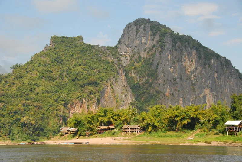 Limestone cliffs rise above riverside stilt houses and long-tail boats on the Mekong near Luang Prabang.