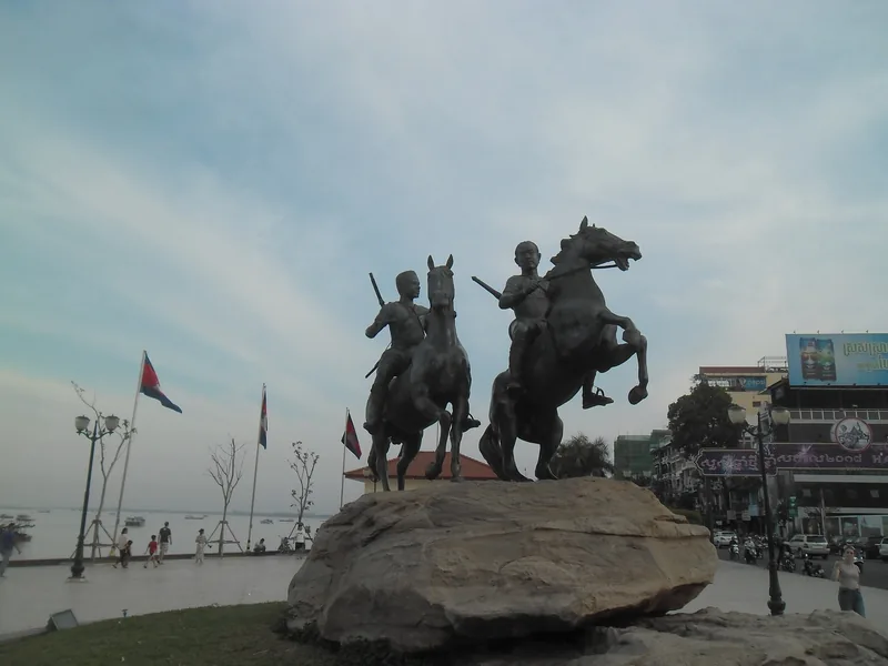 A bronze equestrian statue of two riders on rearing horses stands on a rock along the riverside promenade in Phnom Penh.