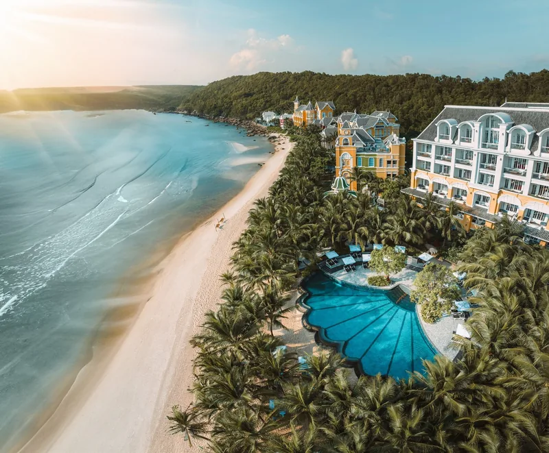 Aerial view of a shell-shaped resort pool, palm trees and beachfront hotel buildings along Phu Quoc's coastline at sunset