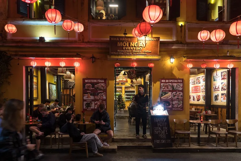 Red silk lanterns glow above a yellow-walled restaurant front with diners seated outside in Hoi An's old town at night.