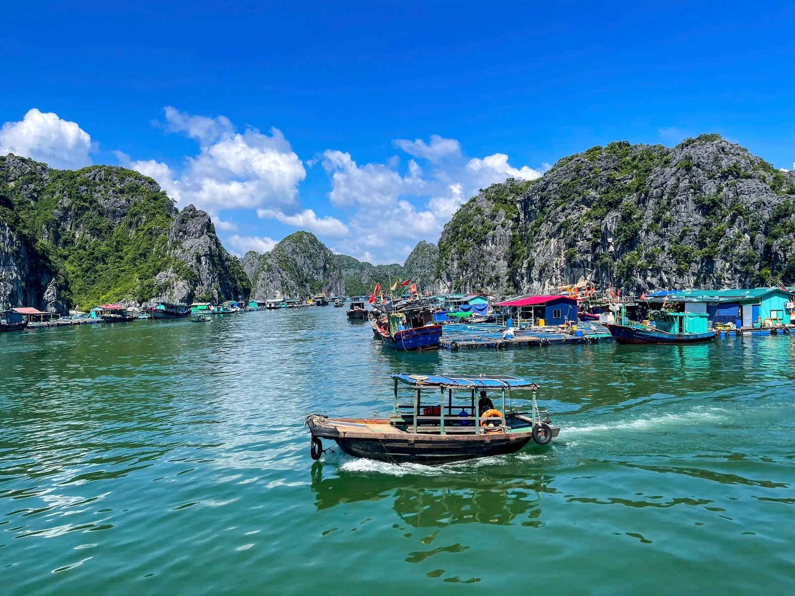 A small wooden boat cruises past a floating fishing village beneath limestone karsts in Lan Ha Bay.